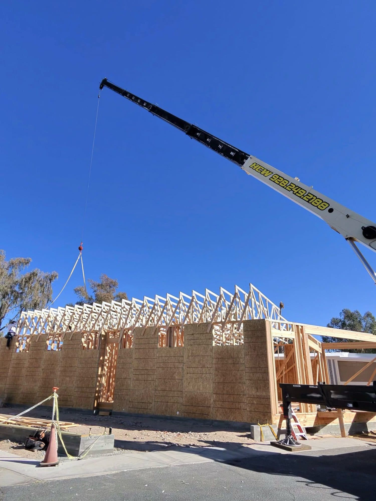 Cream HCW crane boom extended over a wood frame home with roof trusses set and a load on the hook