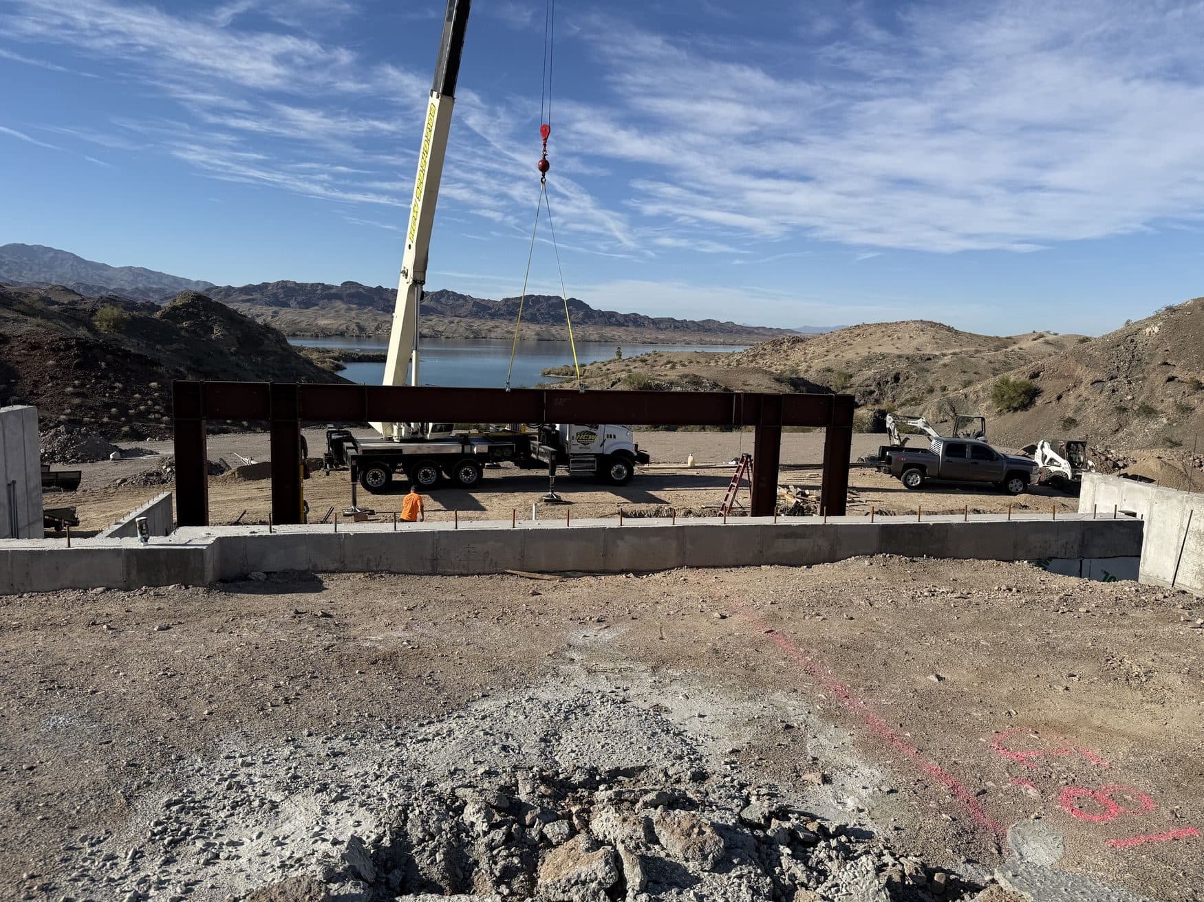 Crane lifting a steel portal frame onto a concrete foundation, with a lake and desert mountains behind
